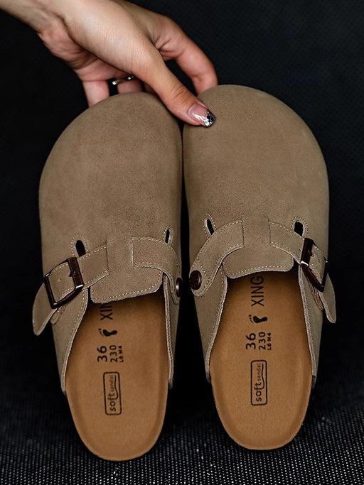 Pair of brown suede clogs with buckles held by a hand on a dark background