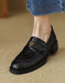 Black loafer shoe with a gold emblem worn with blue jeans on a beige surface