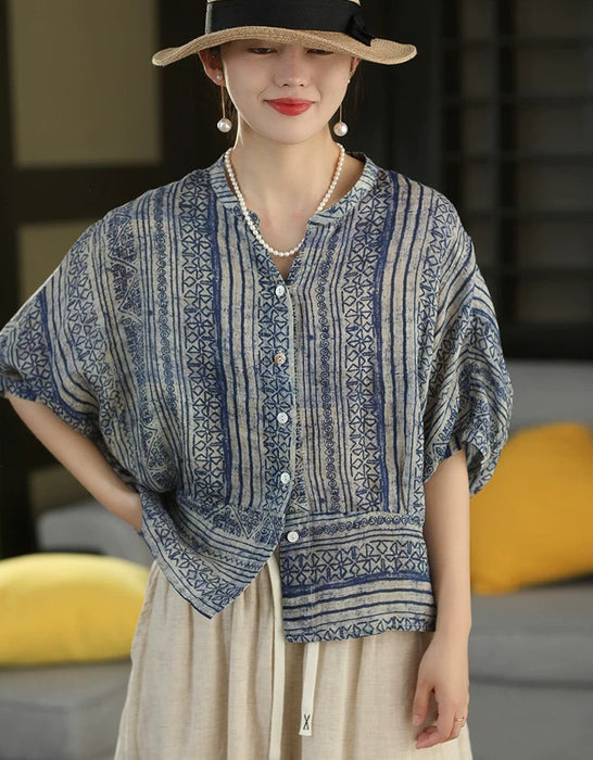 A woman wearing a blue and beige patterned short-sleeve button-up shirt with a relaxed, bohemian style, paired with a beige skirt, pearl necklace, and a black hat with a straw brim.