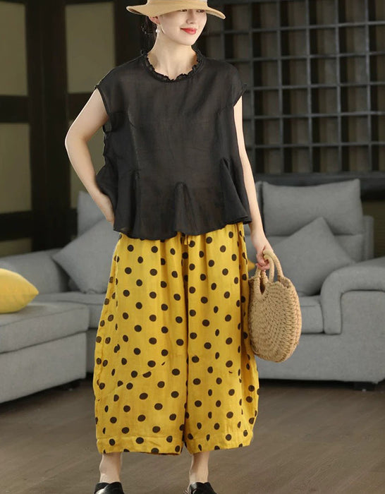 A woman models a two-piece outfit consisting of a black sleeveless ruffled top and yellow wide-leg pants with black polka dots, accessorized with a straw hat and a woven straw bag in a modern living room setting.