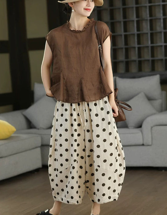 A woman models a two-piece outfit consisting of a brown short-sleeve top with a ruffled neckline and cream-colored wide-leg pants with black polka dots. She is wearing a beige hat and carrying a brown shoulder bag, standing in a living room setting with a gray sofa in the background.