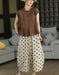 A woman models a two-piece outfit consisting of a brown short-sleeve top with a ruffled neckline and cream-colored wide-leg pants with black polka dots. She is wearing a beige hat and carrying a brown shoulder bag, standing in a living room setting with a gray sofa in the background.